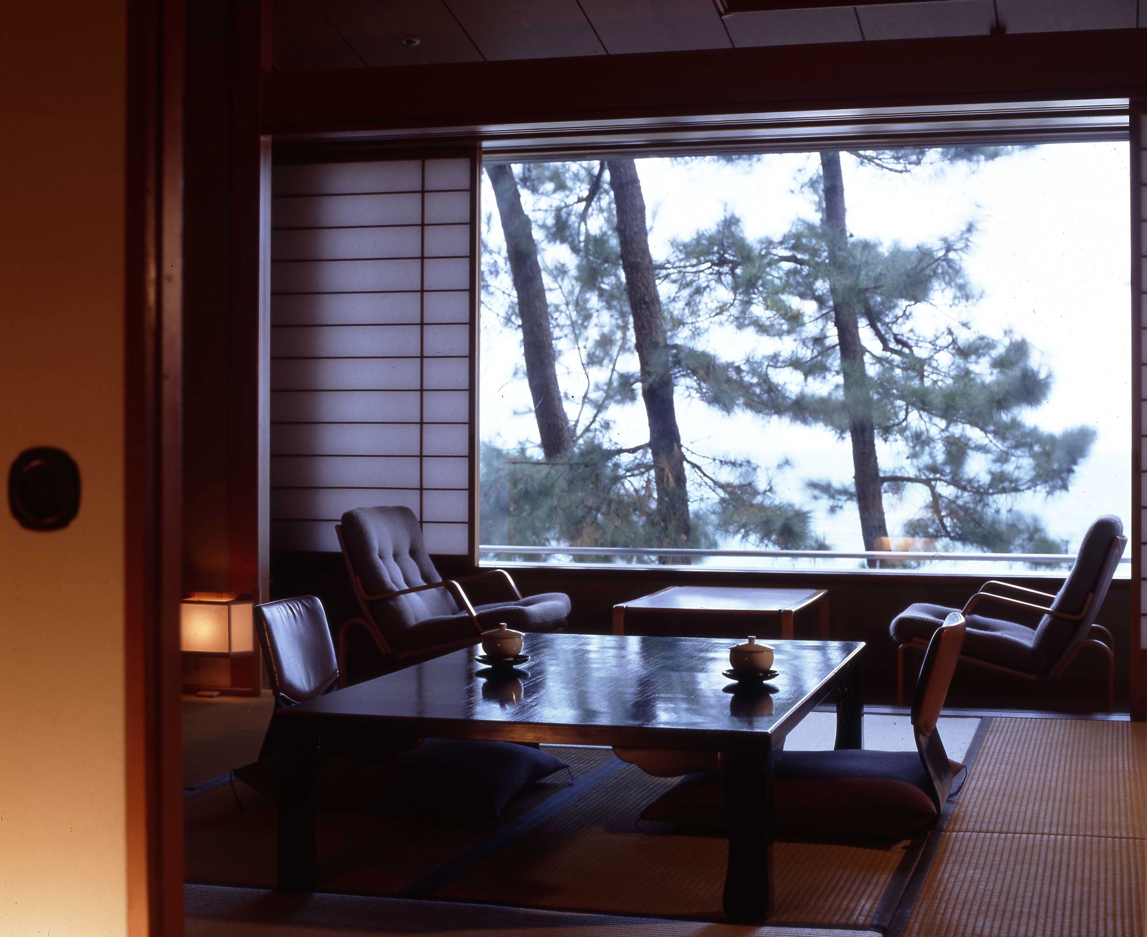 Ein traditioneller japanischer Raum mit Tatami-Bodenbelag, einem niedrigen Tisch mit Tee-Set und Blick auf Kiefern im Freien.