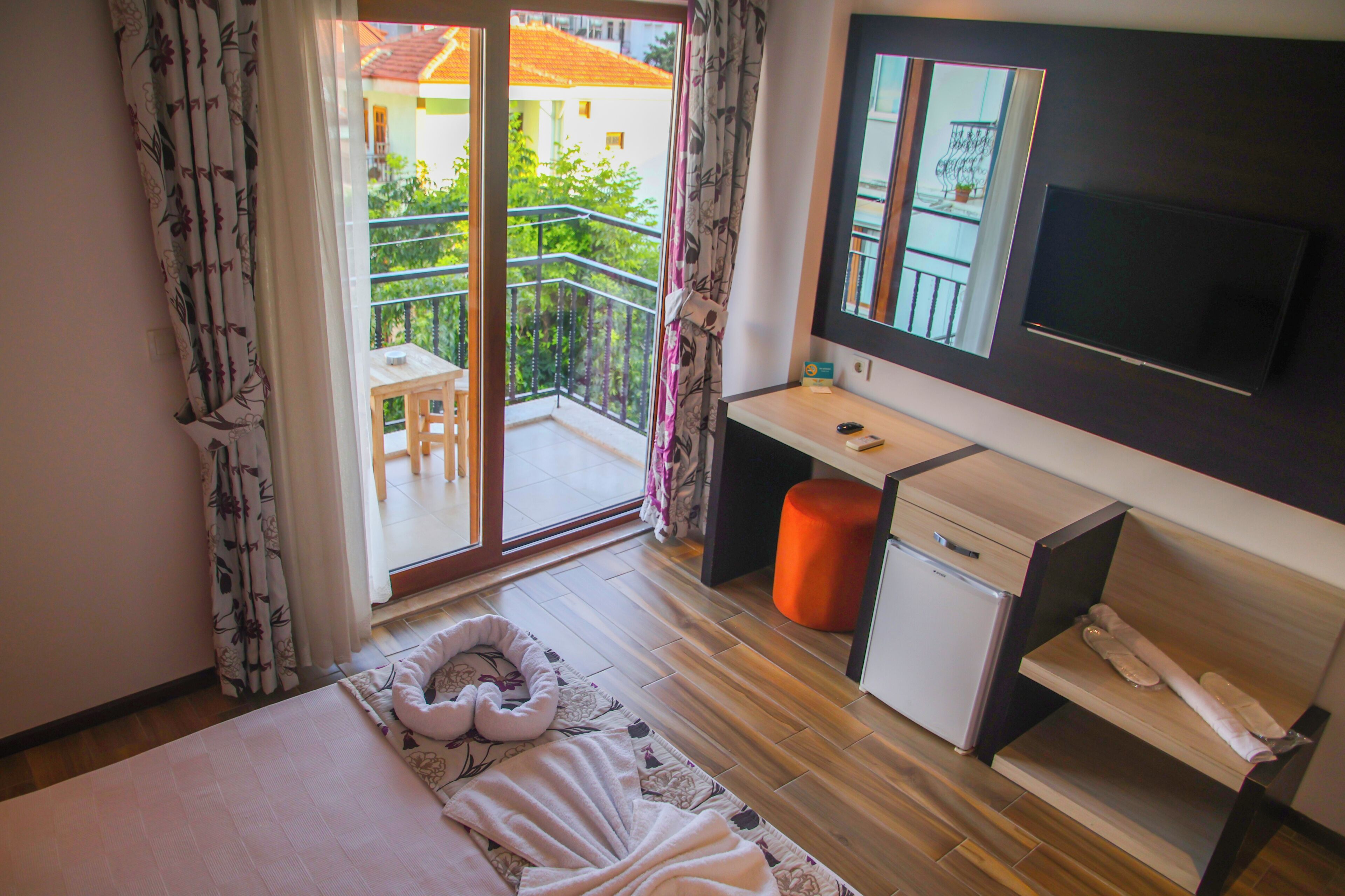 A bedroom with a bed, a desk, a chair, a small table, a TV, and a balcony with a railing.