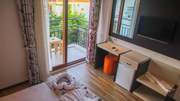 A bedroom with a bed, a desk, a chair, a small table, a TV, and a balcony with a railing.