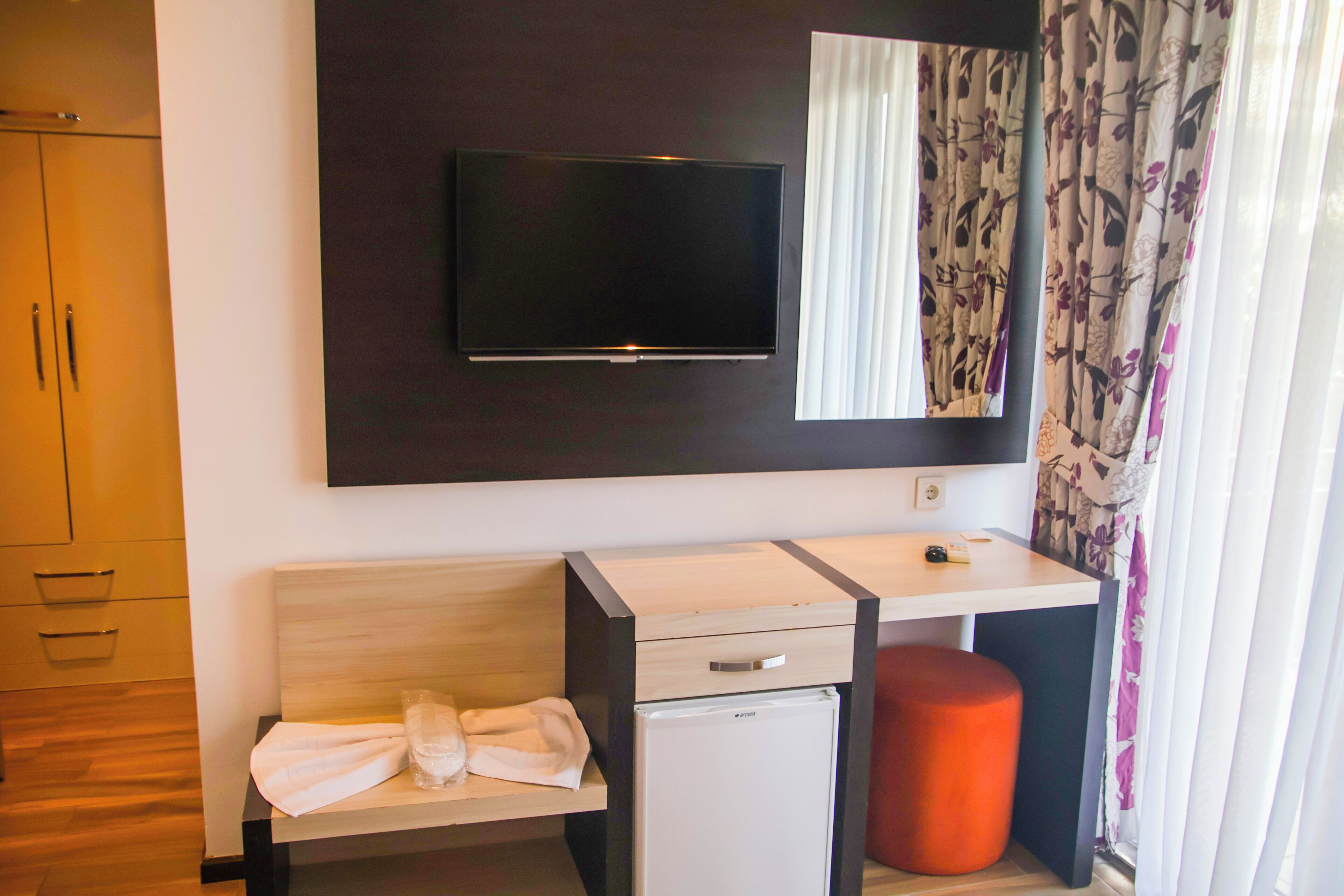 A compact hotel room with a flat-screen TV mounted above a wooden desk, a small refrigerator, a single drawer, and a red ottoman.