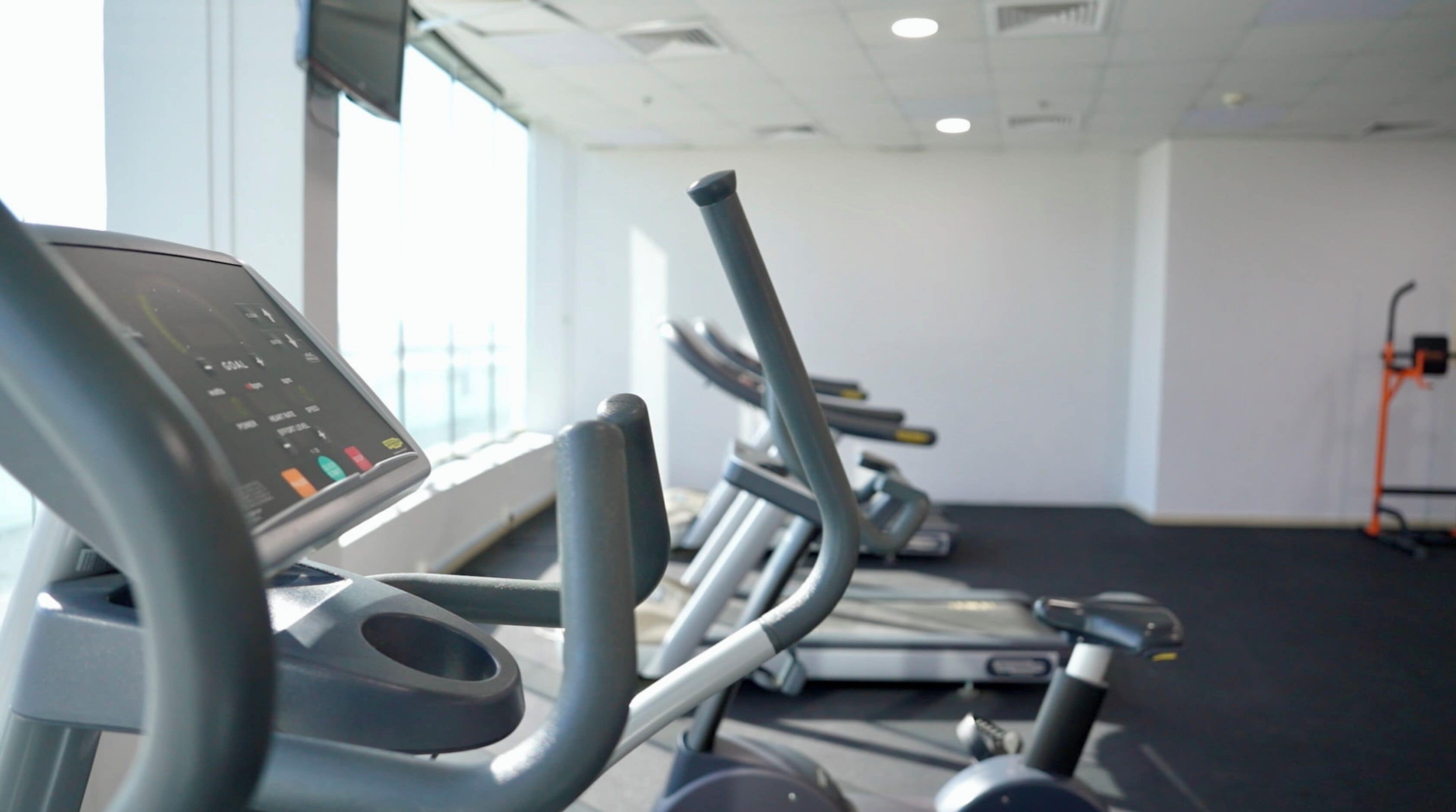 Eine Reihe von stehenden Ergometer mit digitalen Anzeigen, ein Fenster mit Jalousien und eine schlichte Wand mit Steckdose.