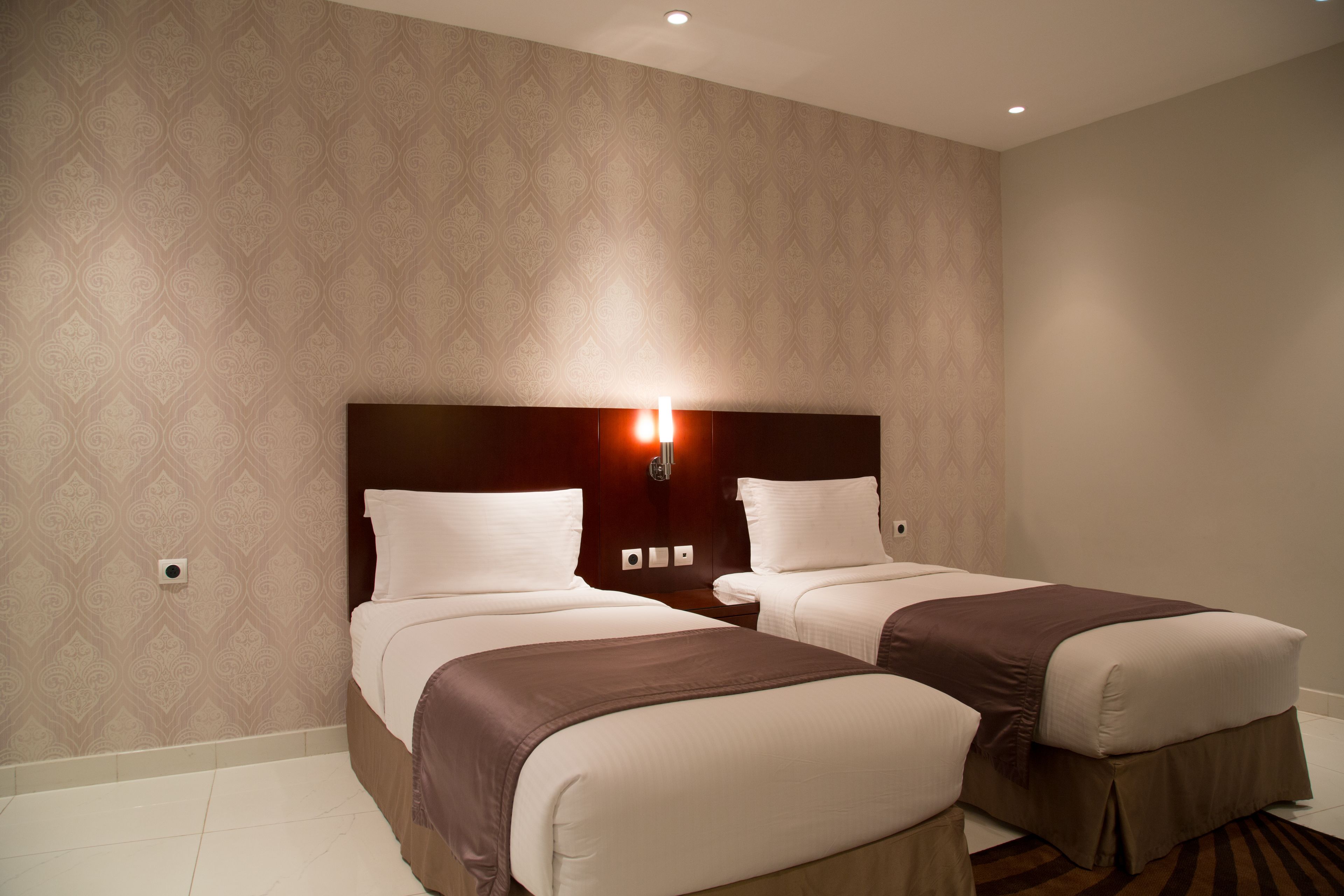 Habitación de hotel con dos camas, cabecero de madera, luz de pared y papel tapiz estampado.