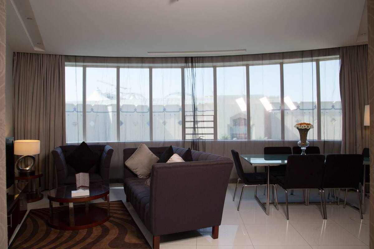 A modern hotel room with a sofa, a small table, a dining area with a table and chairs, and large windows offering a view of the cityscape.