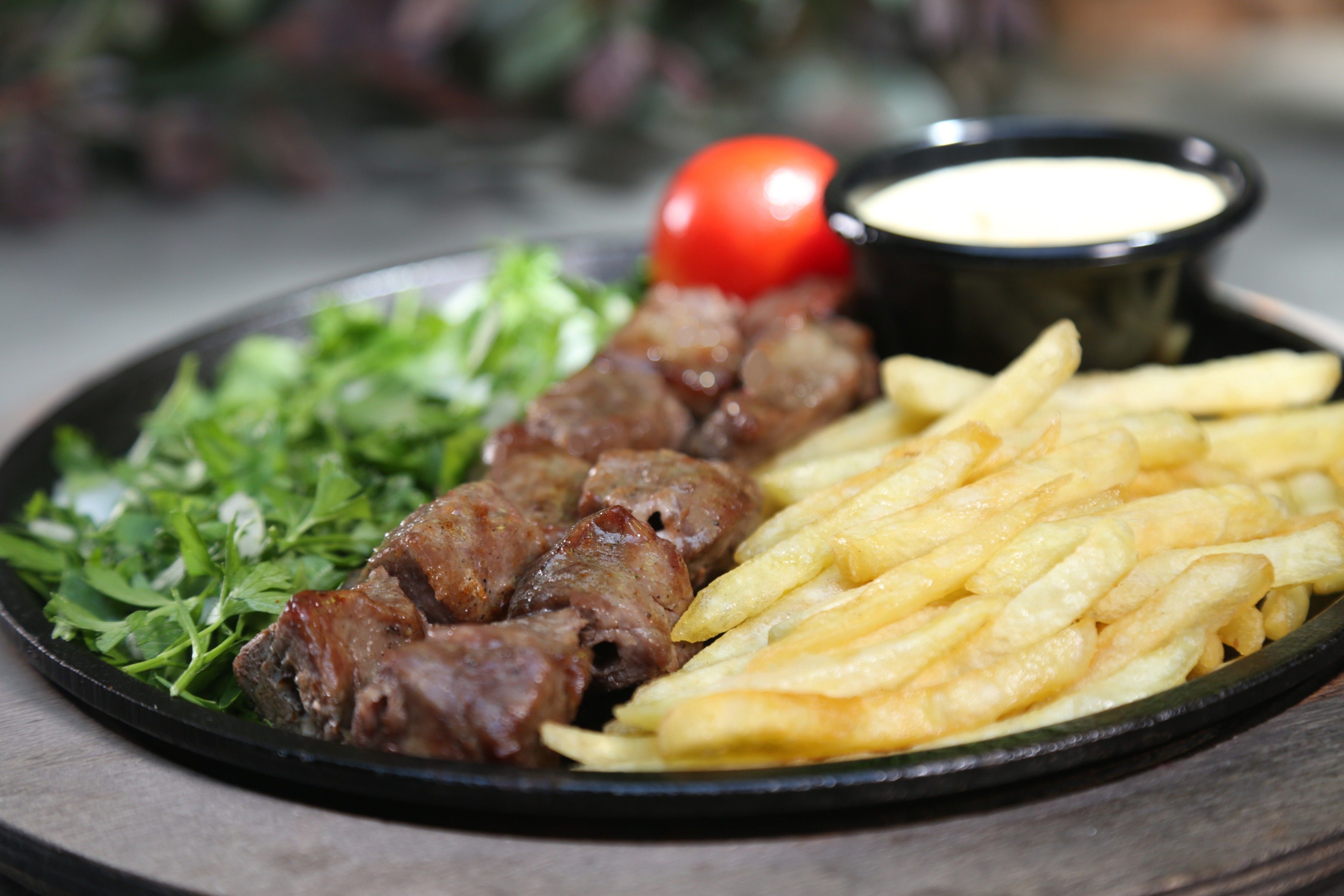 A plate with kebabs, fries, a tomato, and a small bowl of sauce.