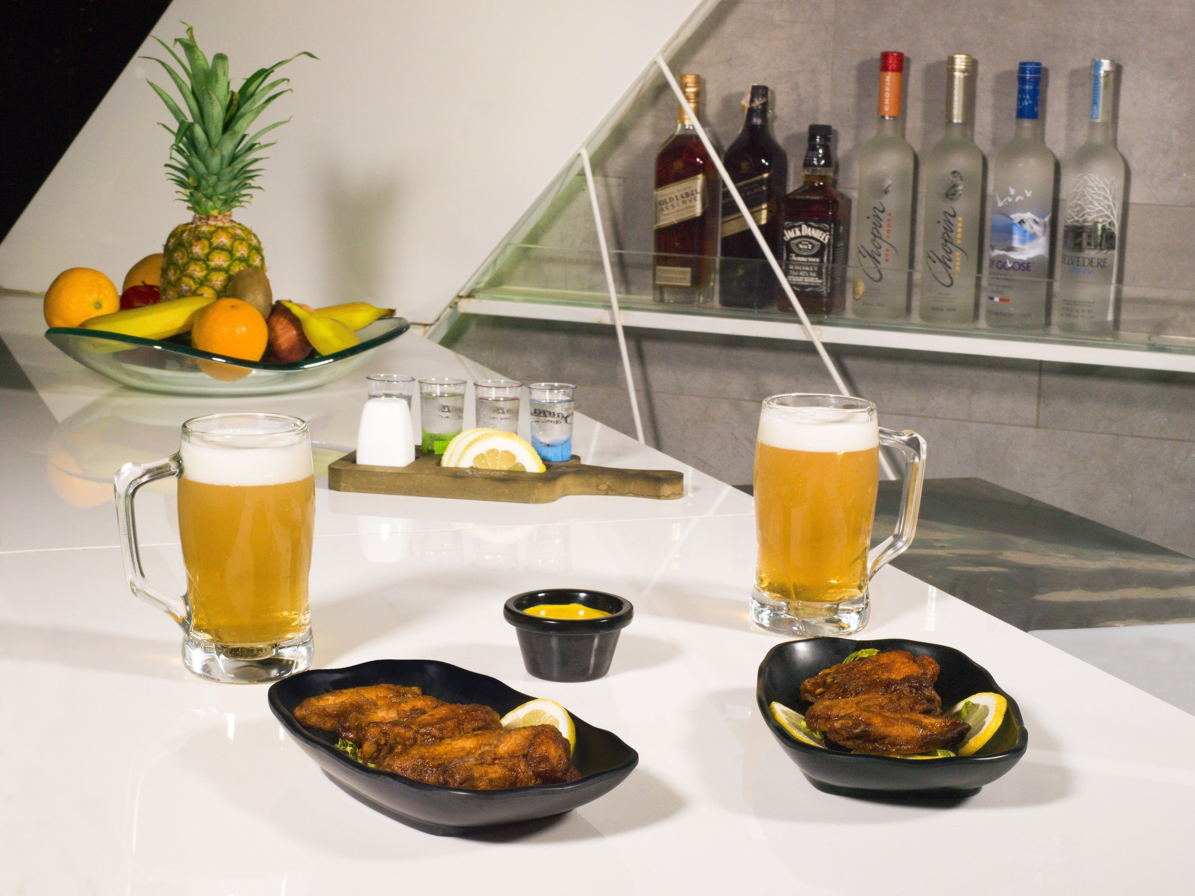 Una zona de bar con dos jarras de cerveza, un bol de fruta y una variedad de botellas de licor.