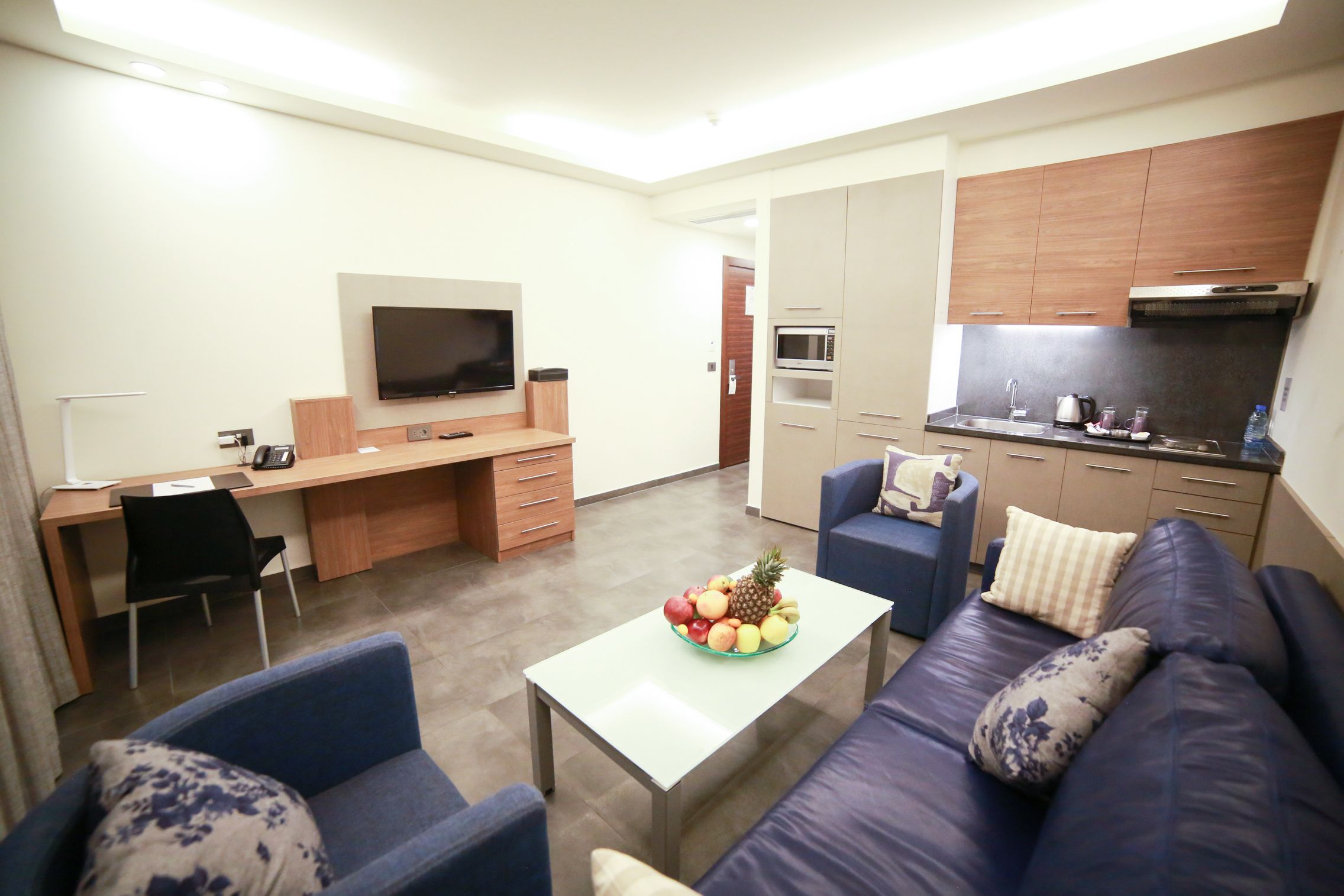 A modern living room with a sofa, coffee table, and a TV. There is a kitchen area with cabinets and appliances.