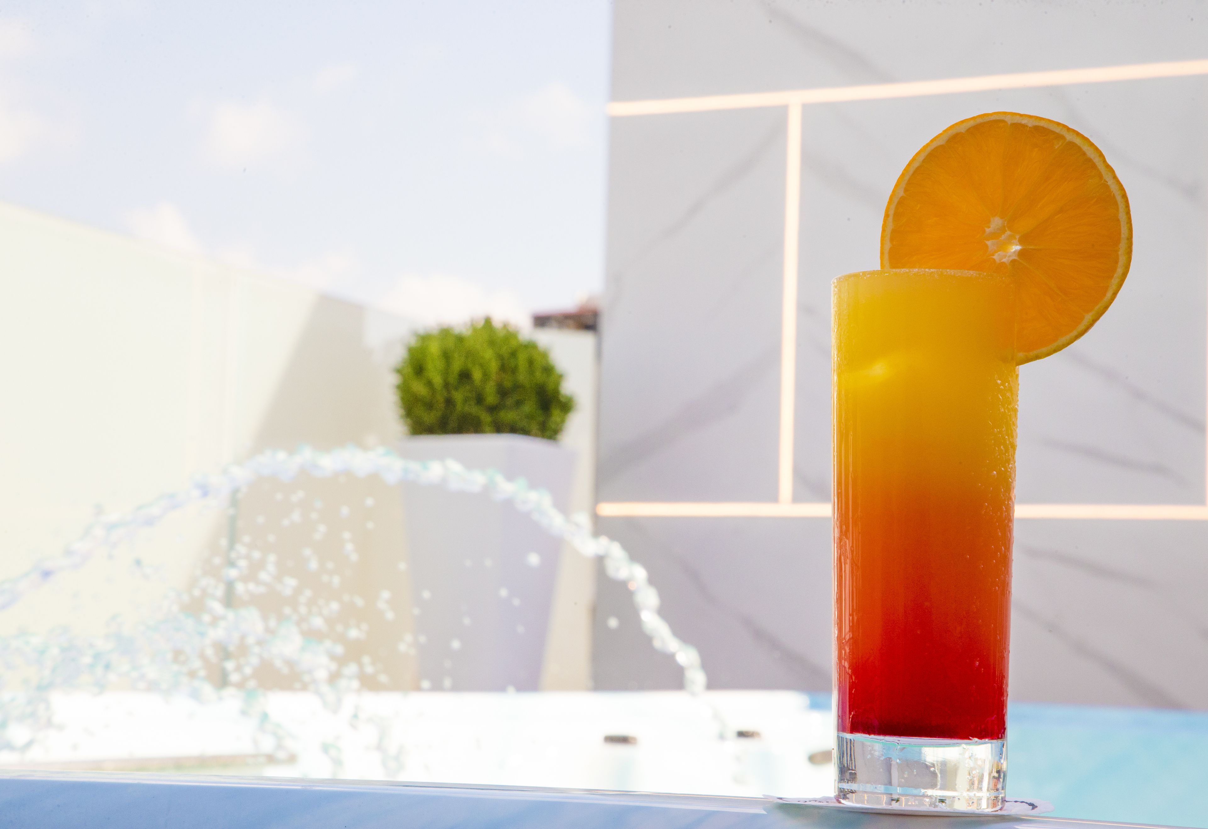 A tall glass with a gradient drink and an orange slice garnish, placed on a table with a water fountain in the background.
