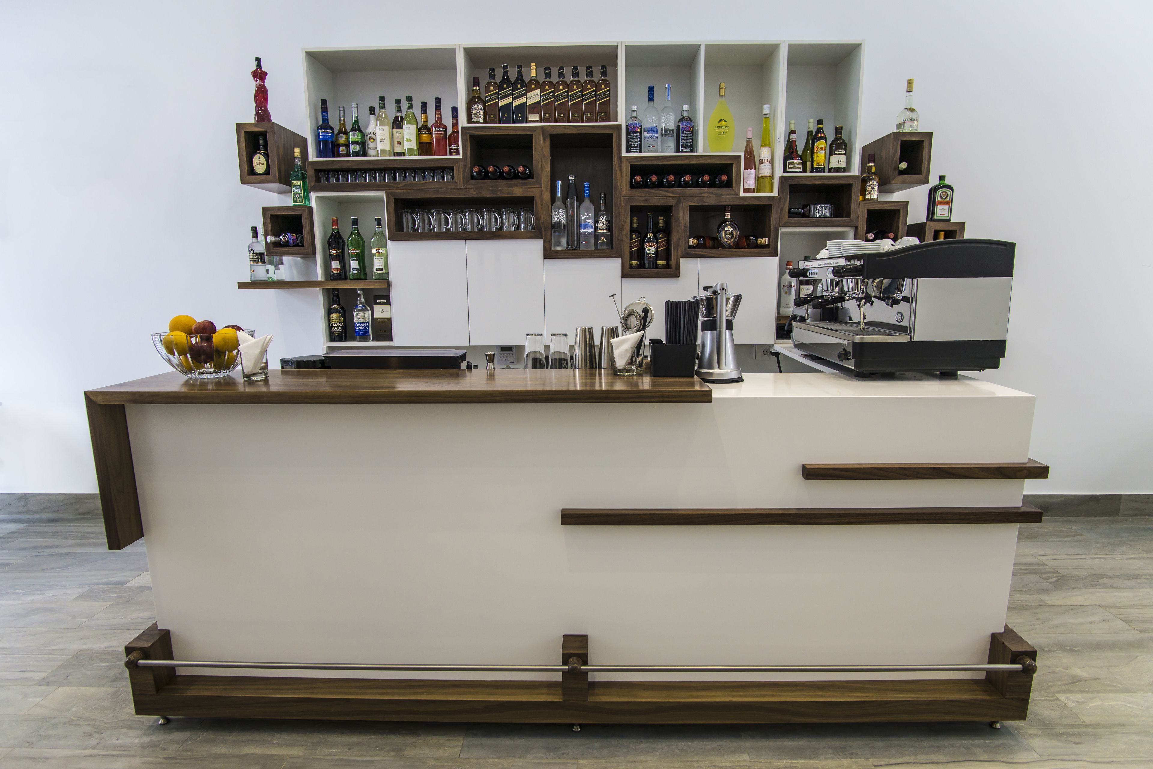 Een bar met een wit aanrecht, houten planken met flessen en een koffieapparaat.