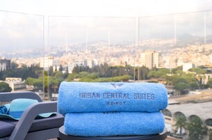 Un balcón con vistas a un paisaje urbano, una toalla azul con el texto "URBAN CENTRAL SUITES BEIRUT" y una silla.