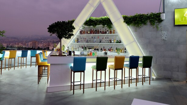 Zona de bar moderna con un original estante triangular que exhibe diferentes botellas, rodeada de taburetes de bar de colores.