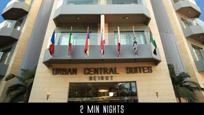 Un hotel llamado "URBAN CENTRAL SUITES BEIRUT" con varias banderas, cielo despejado y un avión en el fondo.