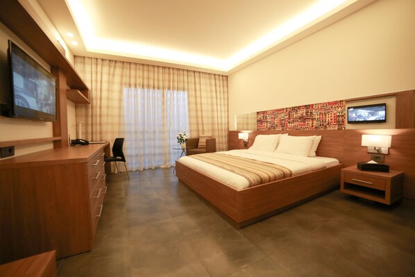Habitación de hotel con una cama grande, un escritorio con cajones, una silla, un televisor montado en la pared y una ventana con cortinas.