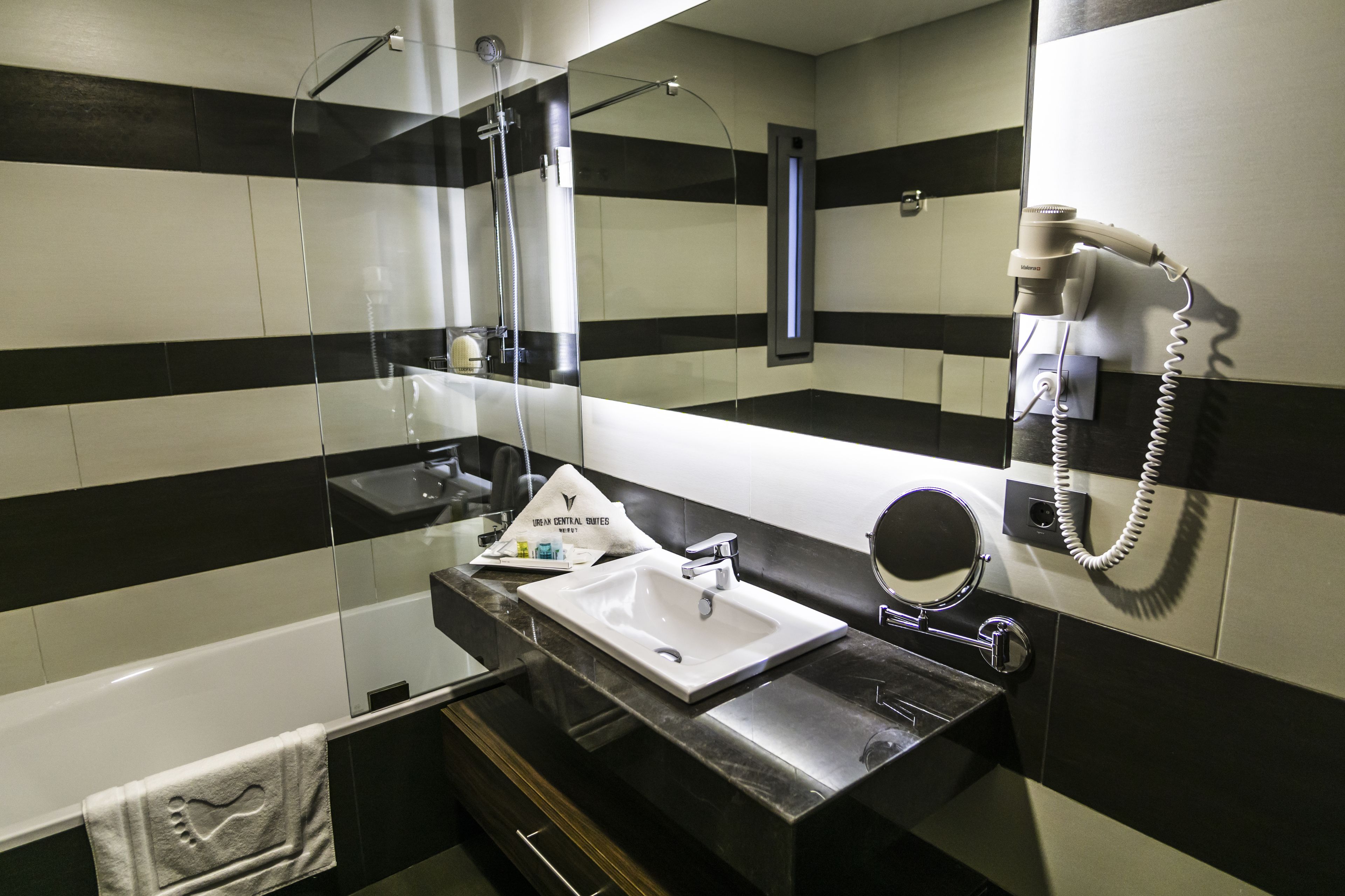 Une salle de bain avec un mur recouvert de carreaux noirs et blancs formant des rayures, un lavabo blanc, un miroir et un sèche-cheveux fixé au mur.