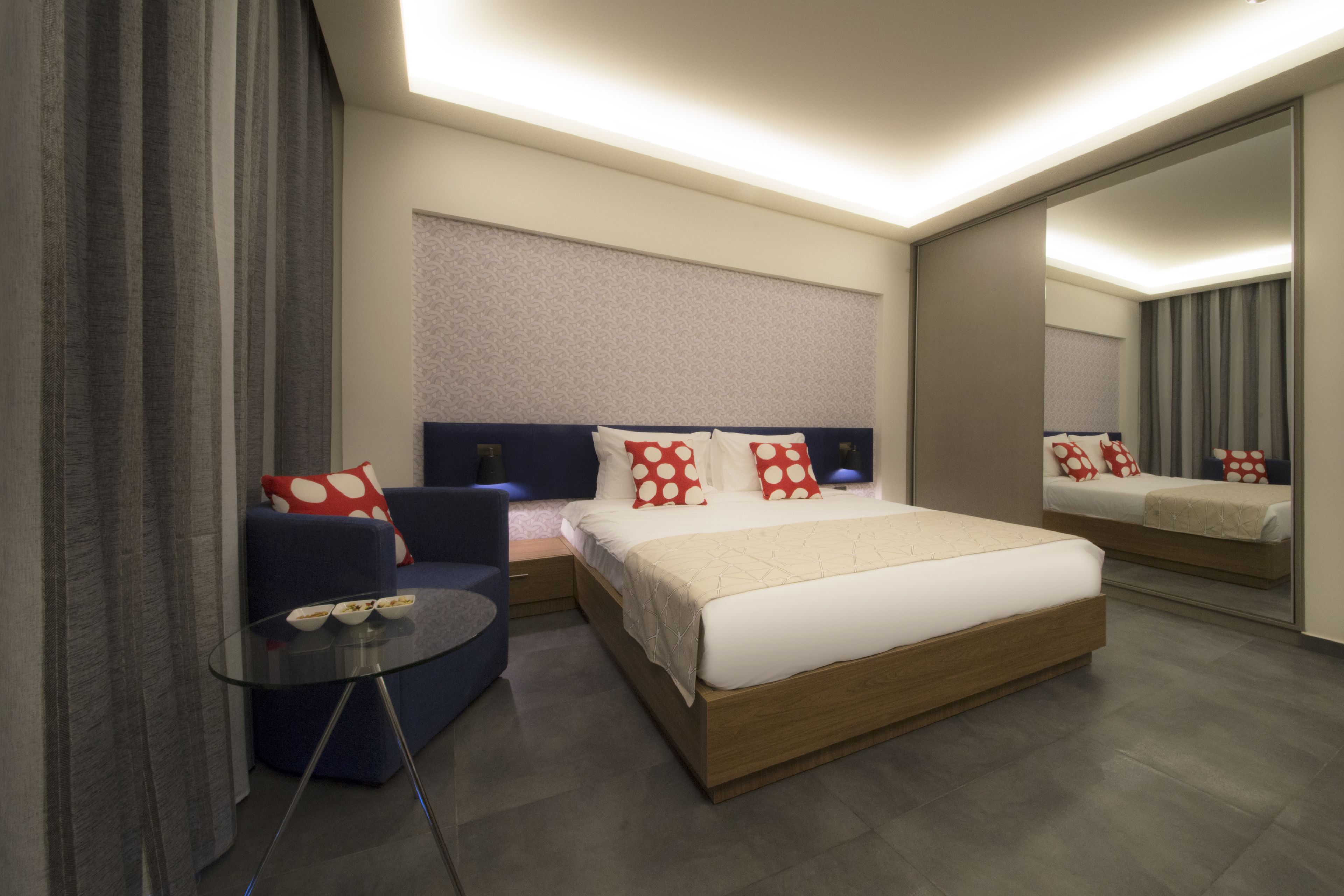 A modern hotel room with a large bed, a seating area with a glass table, and a large mirror.