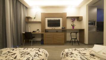 A hotel room with a desk, two chairs, a TV, a small kitchen area, and two beds with floral bedding.