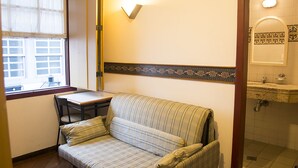 A small room with a sofa, a table, a window, and a bathroom with a sink and mirror. - Hotel Colonial (Ouro Preto)