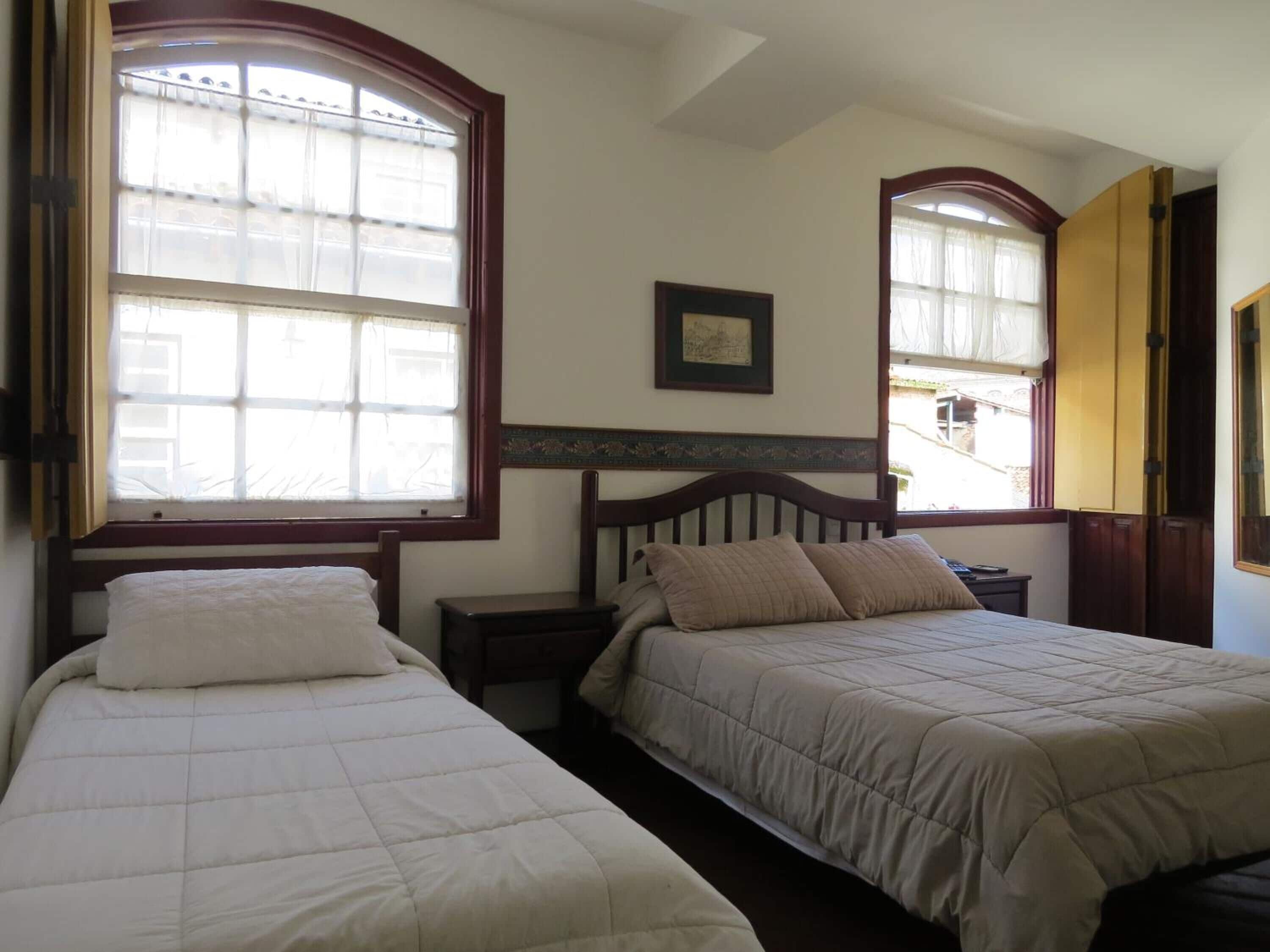 A room with two beds, each with a wooden headboard and a window with curtains. There is a small bedside table with a lamp on each bed.