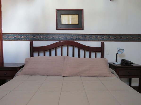 A bedroom with a wooden headboard, a bed with a beige quilt, a nightstand with a lamp, and a framed picture on the wall. - Hotel Colonial (Ouro Preto)