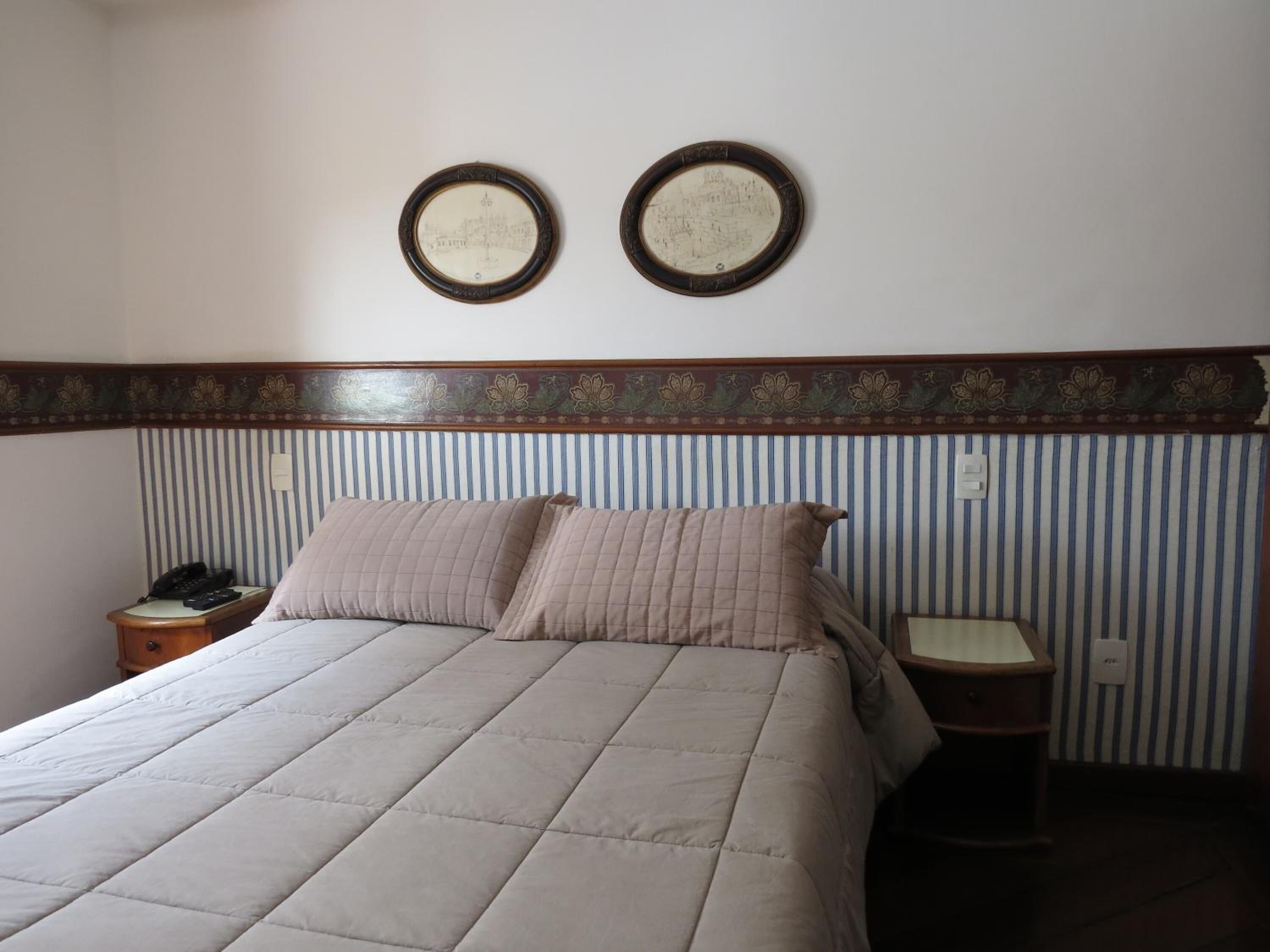 A hotel room with a bed, two bedside tables, a telephone, and two framed pictures on the wall.