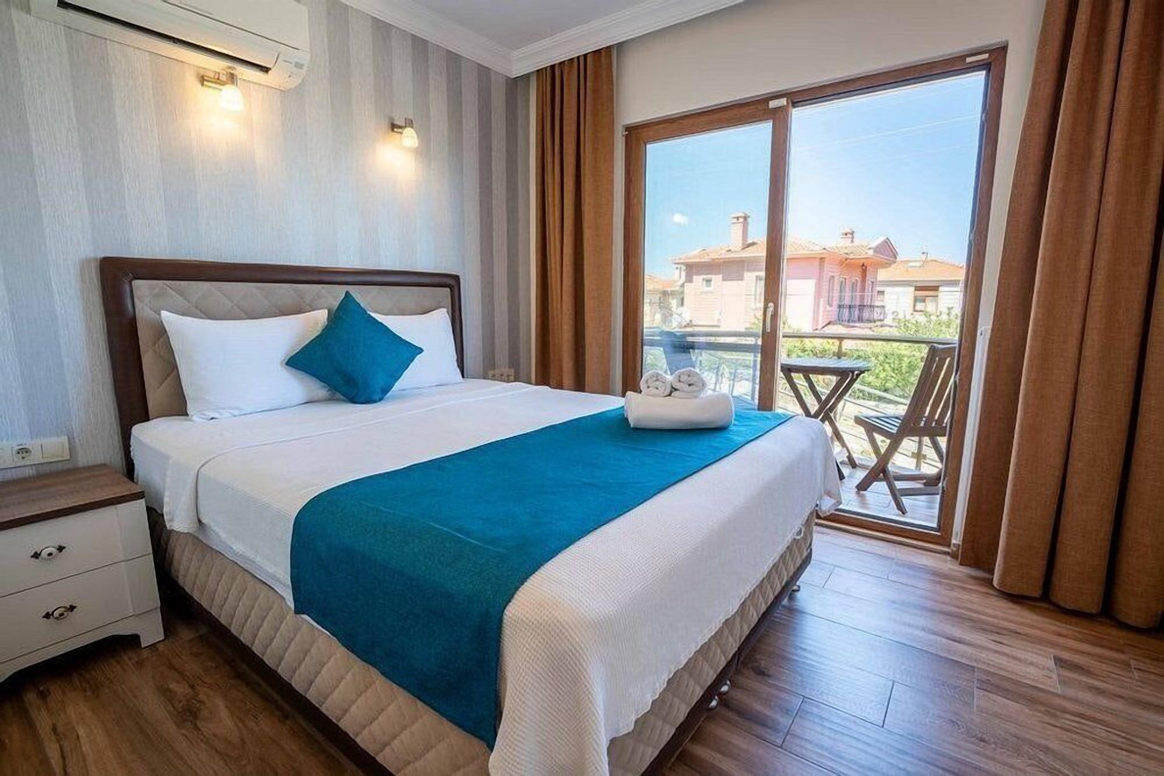 A hotel room with a bed, a nightstand, a table with chairs outside, and a view of buildings.