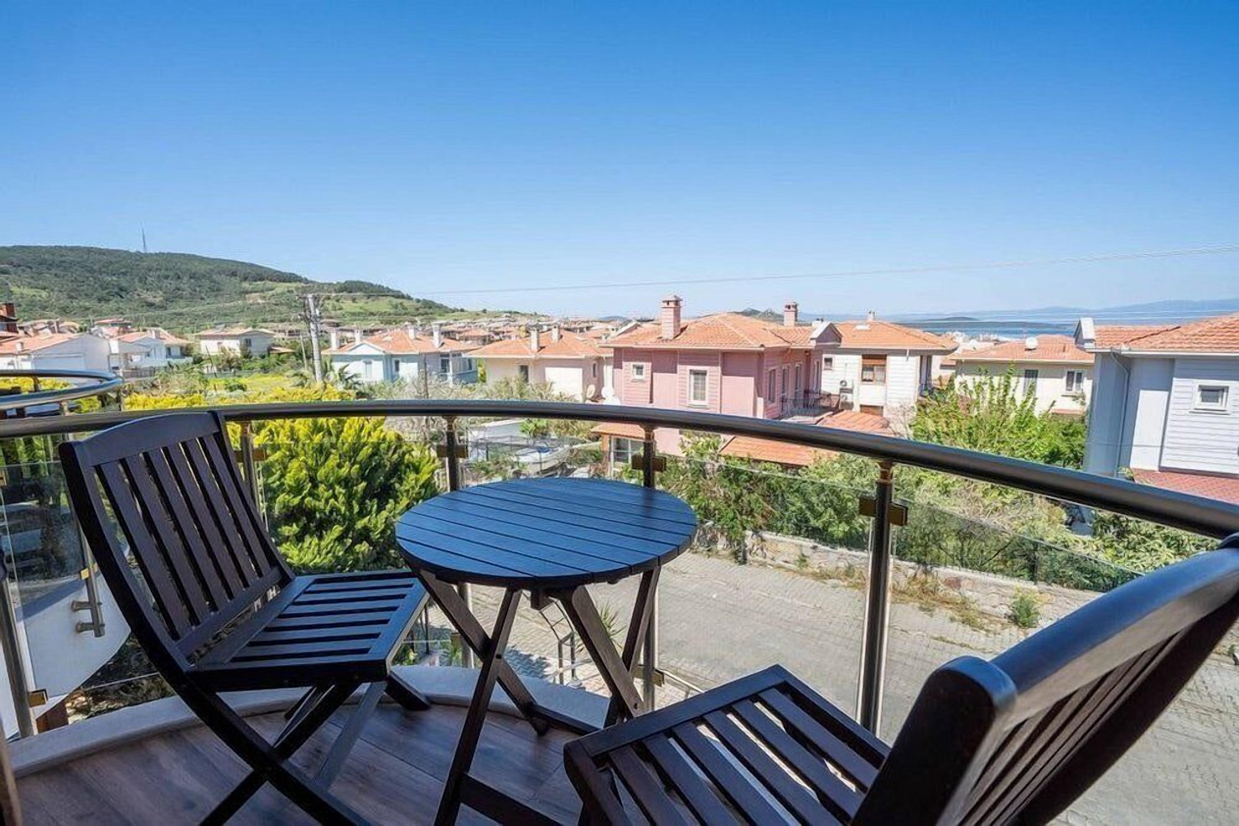 A balcony with two wooden chairs and a small round table, overlooking a residential area with houses and a hilly landscape.