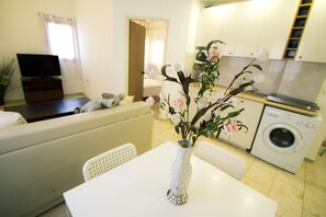 A modern living room with a white sofa, a dining table with a white vase, a television, and a washing machine in the background. - Apartment - Tel Aviv (Tel Aviv)