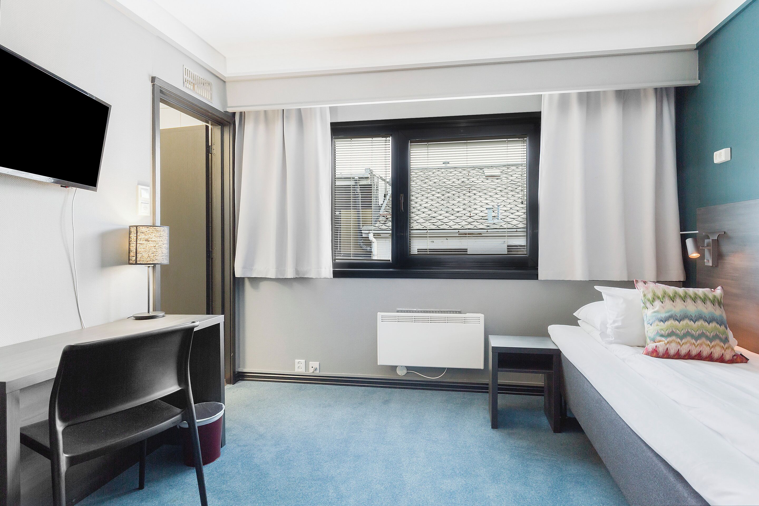 Una habitación de hotel con cama, escritorio, silla, televisor, ventana con cortinas y radiador.