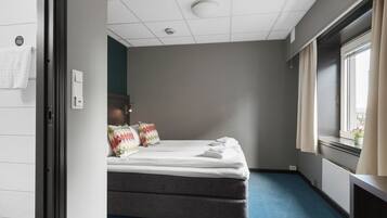 A hotel room with a bed, a window with curtains, a radiator, and a wall-mounted light switch.