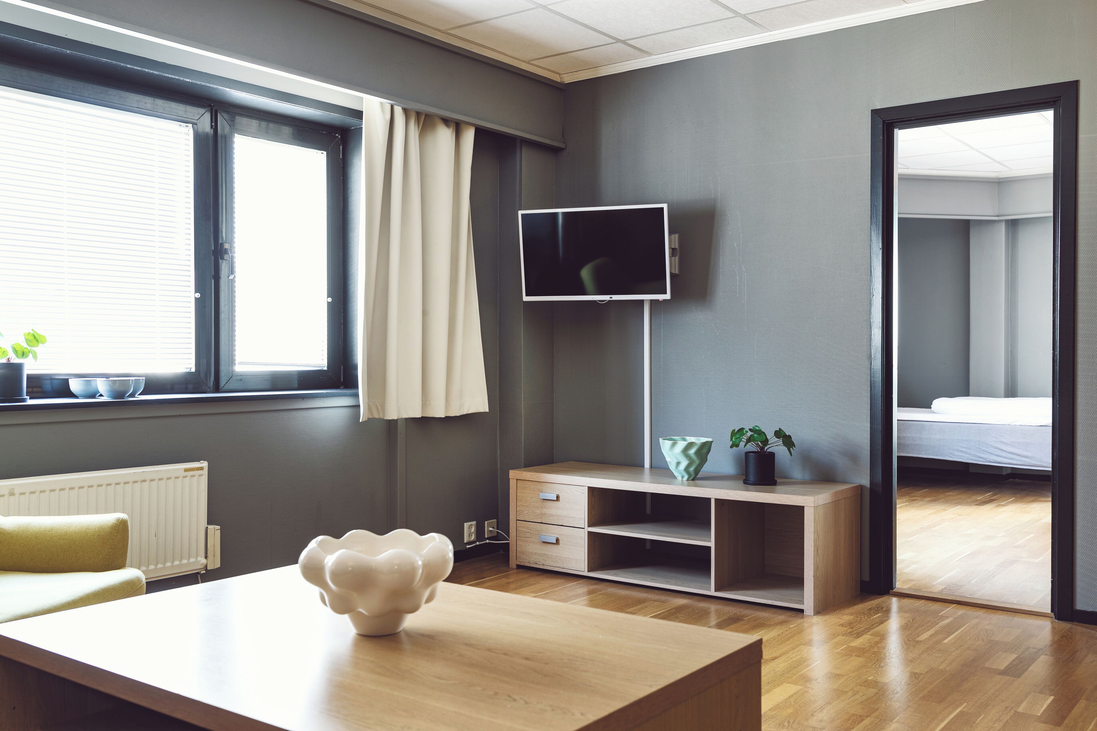 A modern room with a wooden desk, a TV mounted on the wall, a bed in the adjacent room, and a window with curtains.
