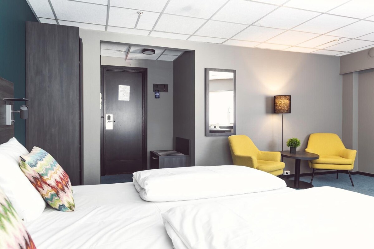 A hotel room with a bed, two yellow chairs, a small table, a mirror, and a ceiling light.
