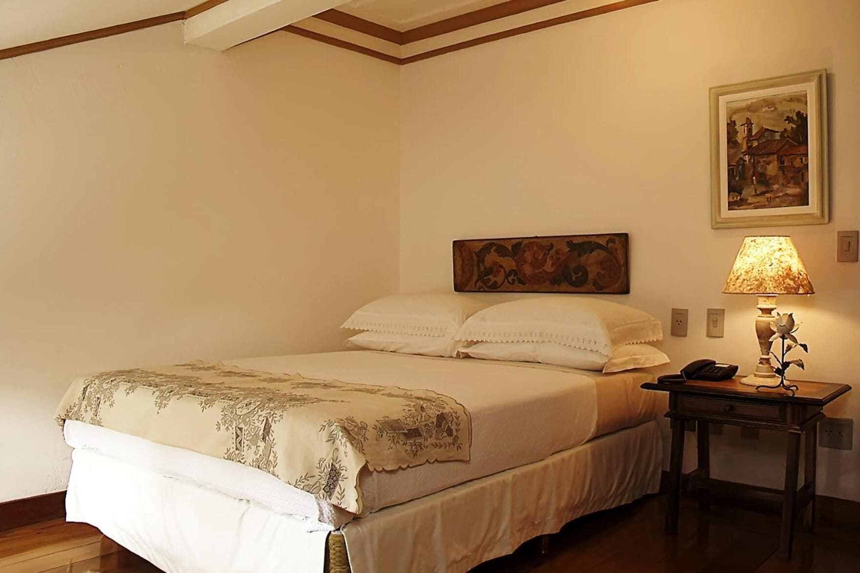 A neatly made bed with a floral bedspread, a bedside table with a lamp, and a painting on the wall.