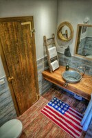 Un baño con puerta de madera, toallero, lavabo con pileta, espejo y alfombra con la bandera estadounidense.