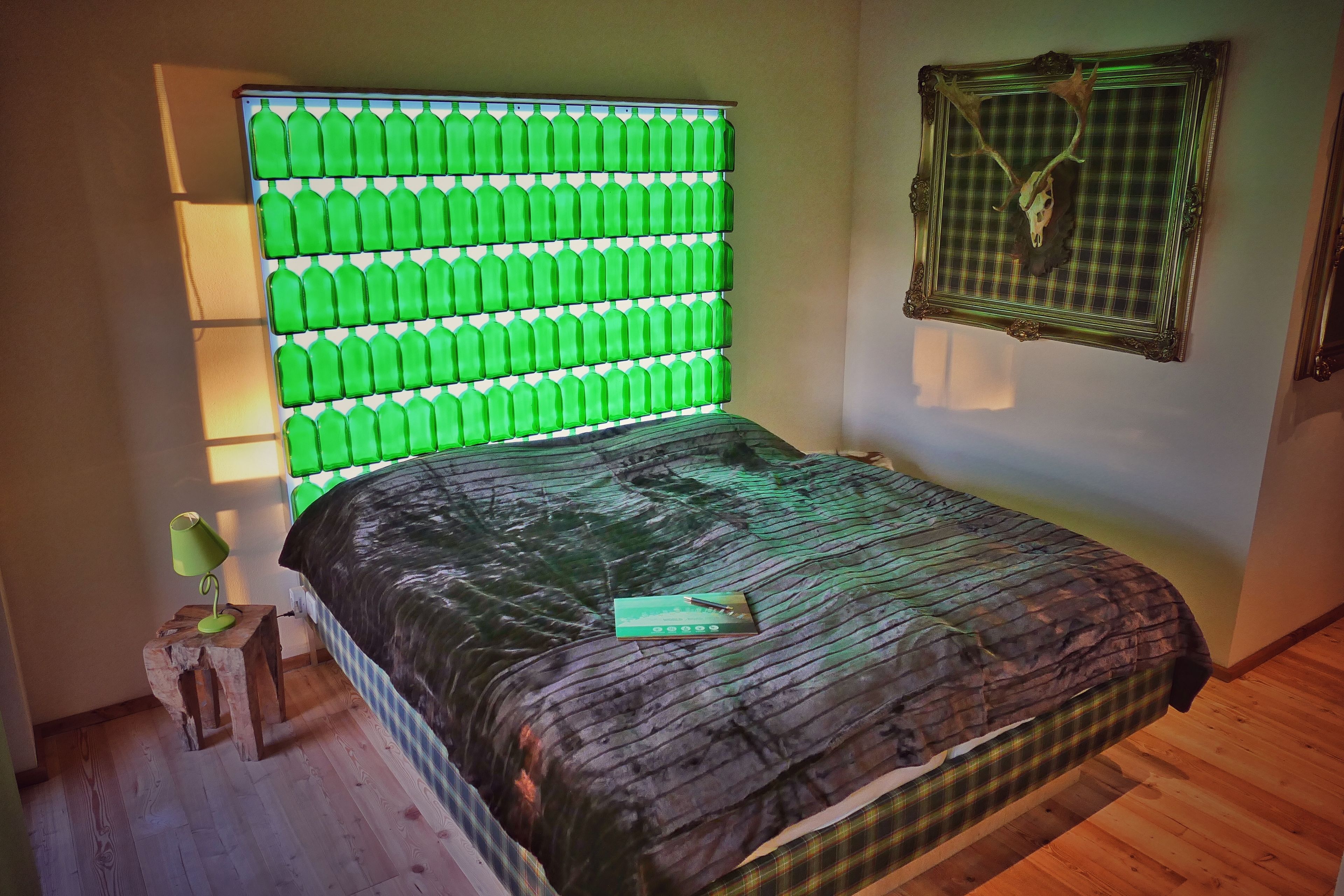 Ein Schlafzimmer mit einer einzigartigen Kopfstütze aus grüner Flasche, einer Bettdecke mit Karomuster und einem gerahmten Damhirschschädel an der Wand.