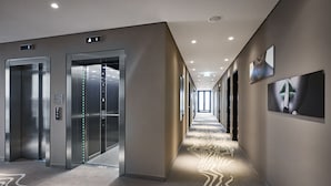 A modern hallway with two elevators, a patterned carpet, and framed artwork on the walls. - LEGERE HOTEL Bielefeld (Bielefeld)