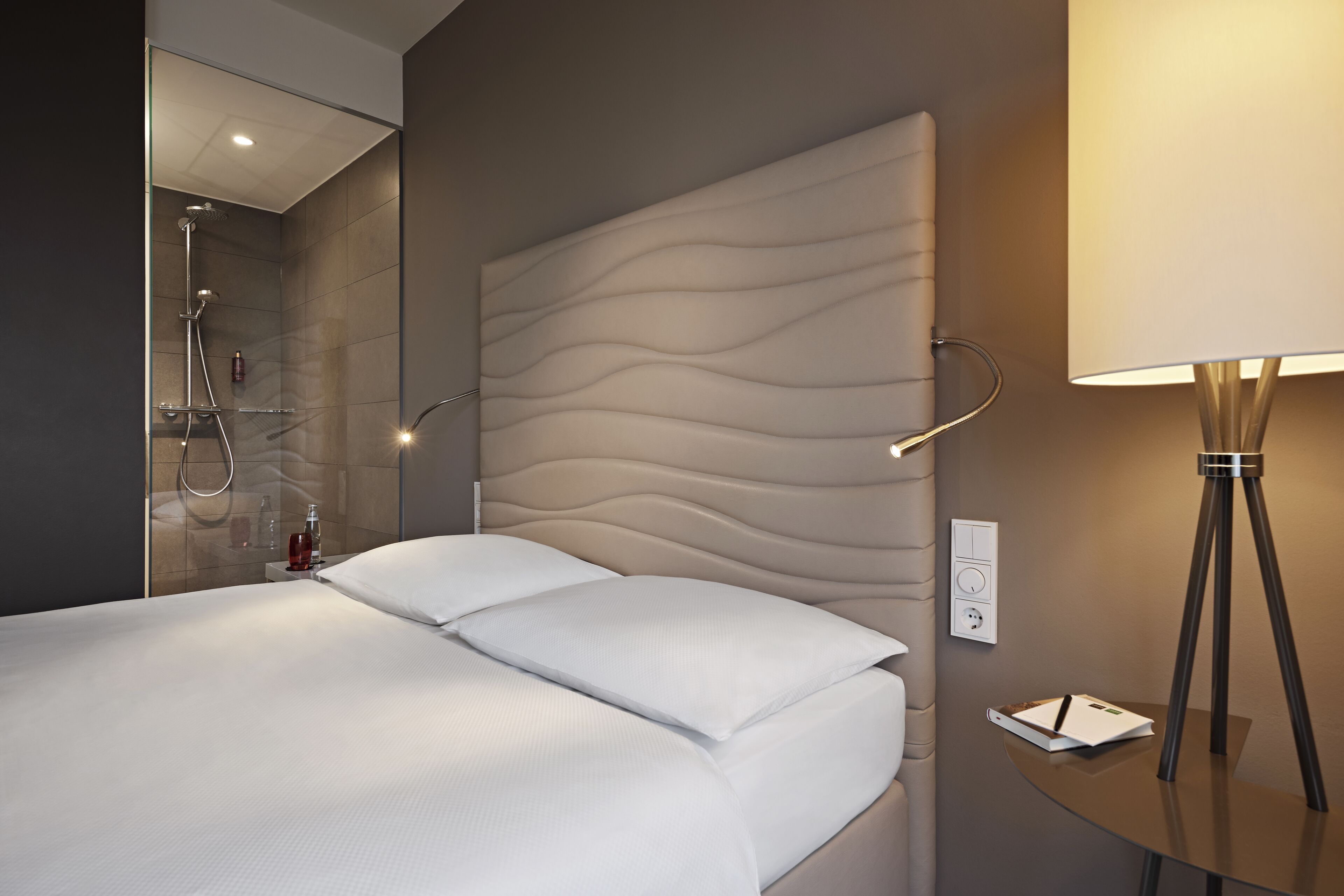 A modern hotel room with a large bed, a bedside table with a lamp, a glass shower enclosure, and a wall-mounted control panel.