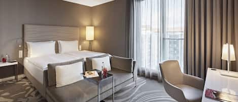 Habitación de hotel con una cama grande, un sofá, un escritorio y una silla. Tiene una ventana con cortinas y una lámpara en el escritorio.