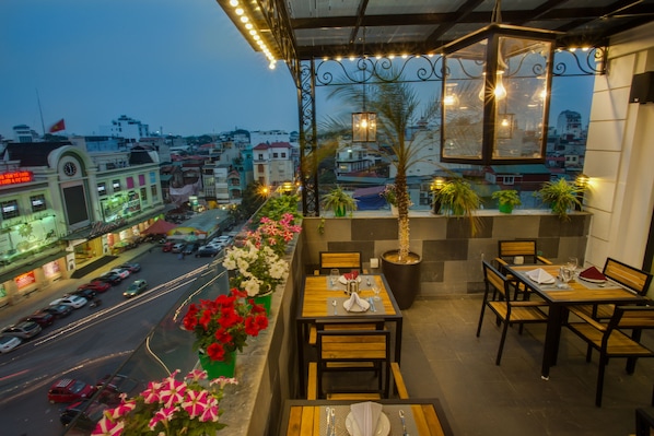 Un área de comedor al aire libre con mesas y sillas, plantas en macetas y vista panorámica de la ciudad.