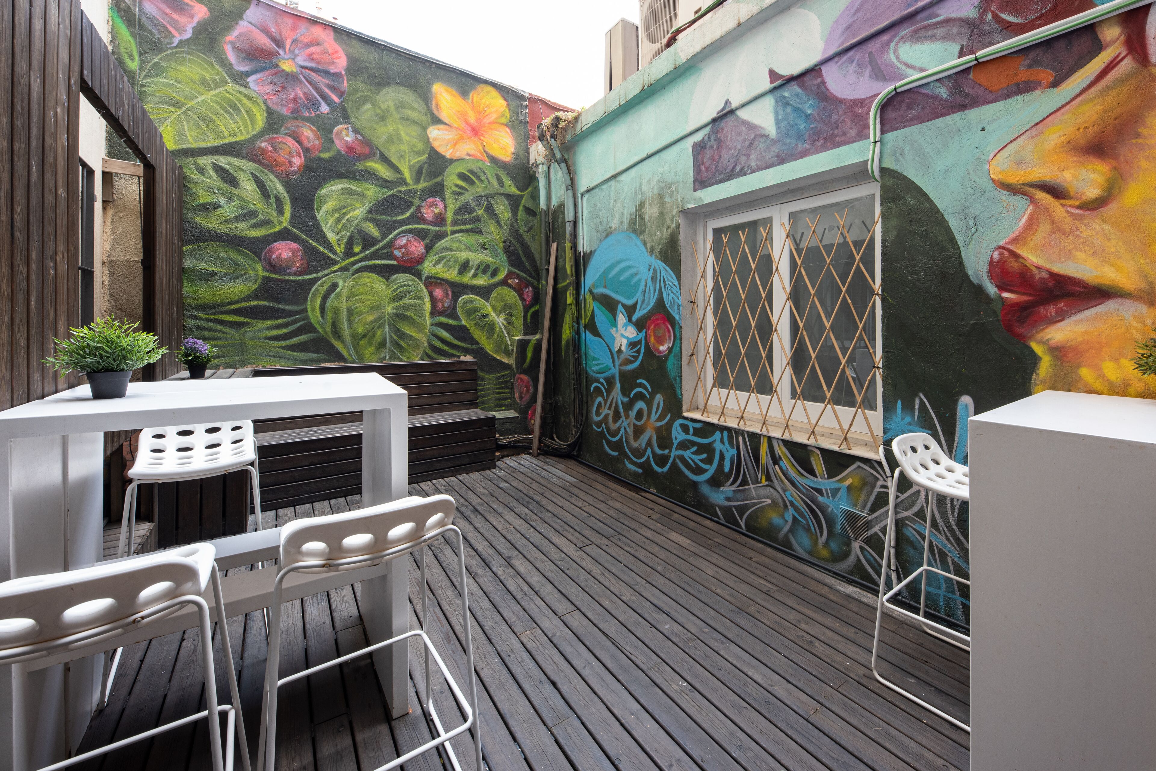 An outdoor patio with a wooden deck, white metal chairs, a table, and a potted plant.