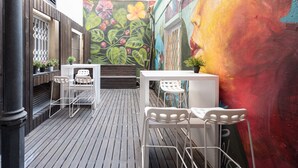 Un patio exterior estrecho con piso de madera, sillas de metal blanco y una mesa blanca. Un mural colorido con diseños florales y abstractos adorna la pared.