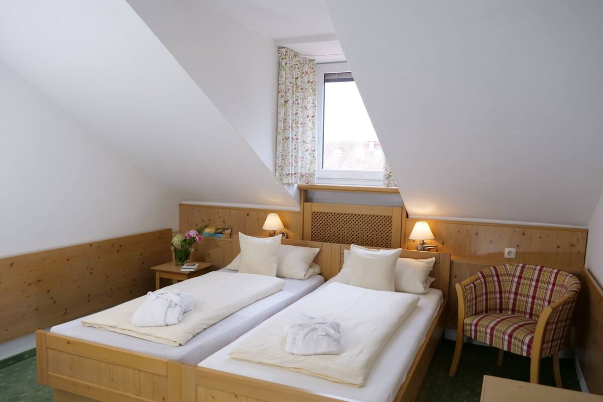 A room with two wooden beds, a small table, a chair, and a window with floral curtains.
