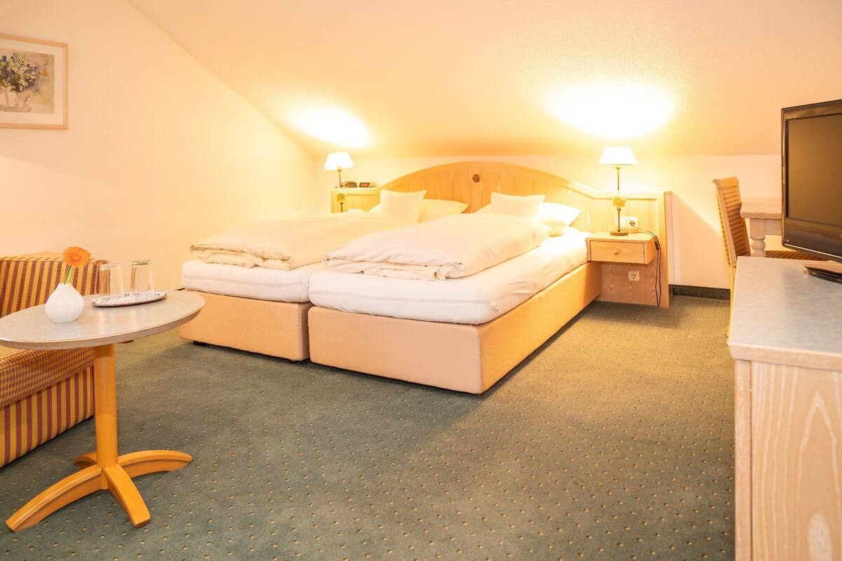 A hotel room with a double bed, two bedside tables with lamps, a small round table with a vase, a television, and a chair.