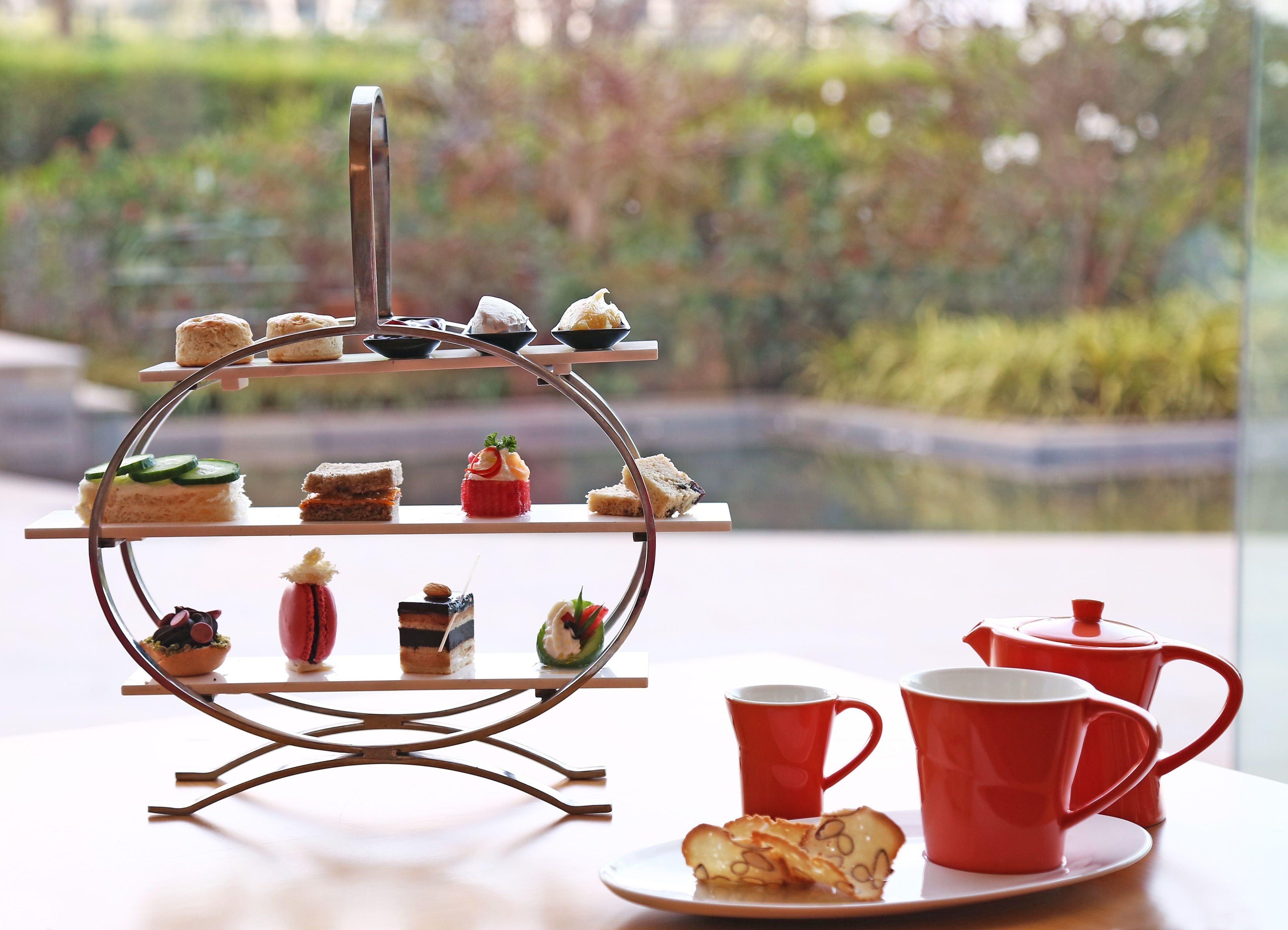 A tiered dessert stand with various pastries and a tea set on a table by a window with a view of greenery.