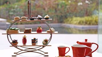 A tiered dessert stand with various pastries and a tea set on a table by a window with a view of greenery.