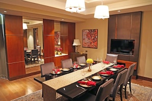 A dining area with a long table set for six, a television mounted on the wall, and a mirror reflecting the room. - Taj Bangalore (Devanahalli)