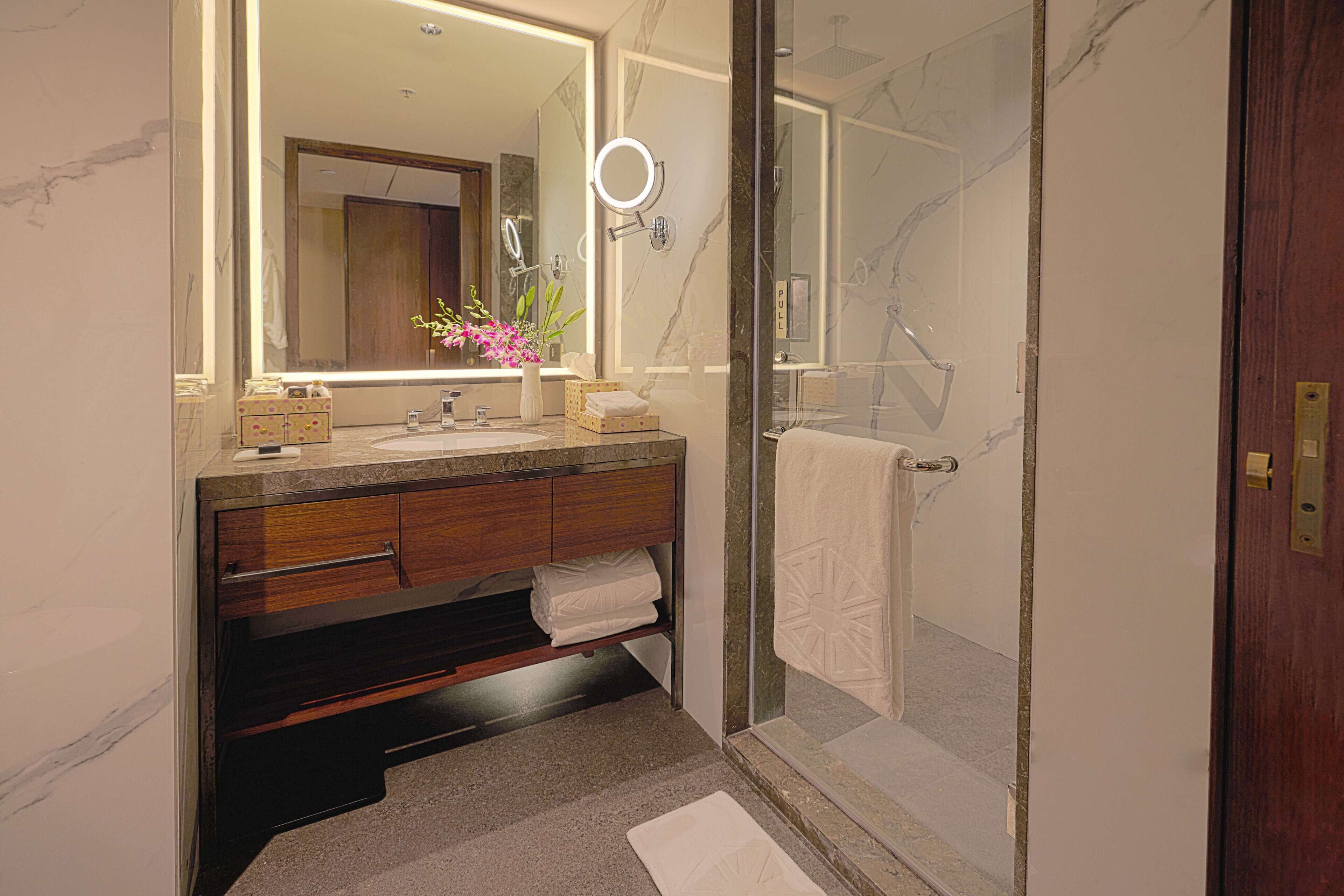 Un baño moderno con tocador de mármol, un espejo grande con luz de aro, cabina de ducha de vidrio y una puerta de madera.