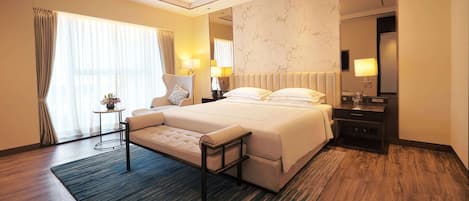 A modern hotel room with a large bed, bedside tables, a small side table with a lamp, and a large window with curtains.