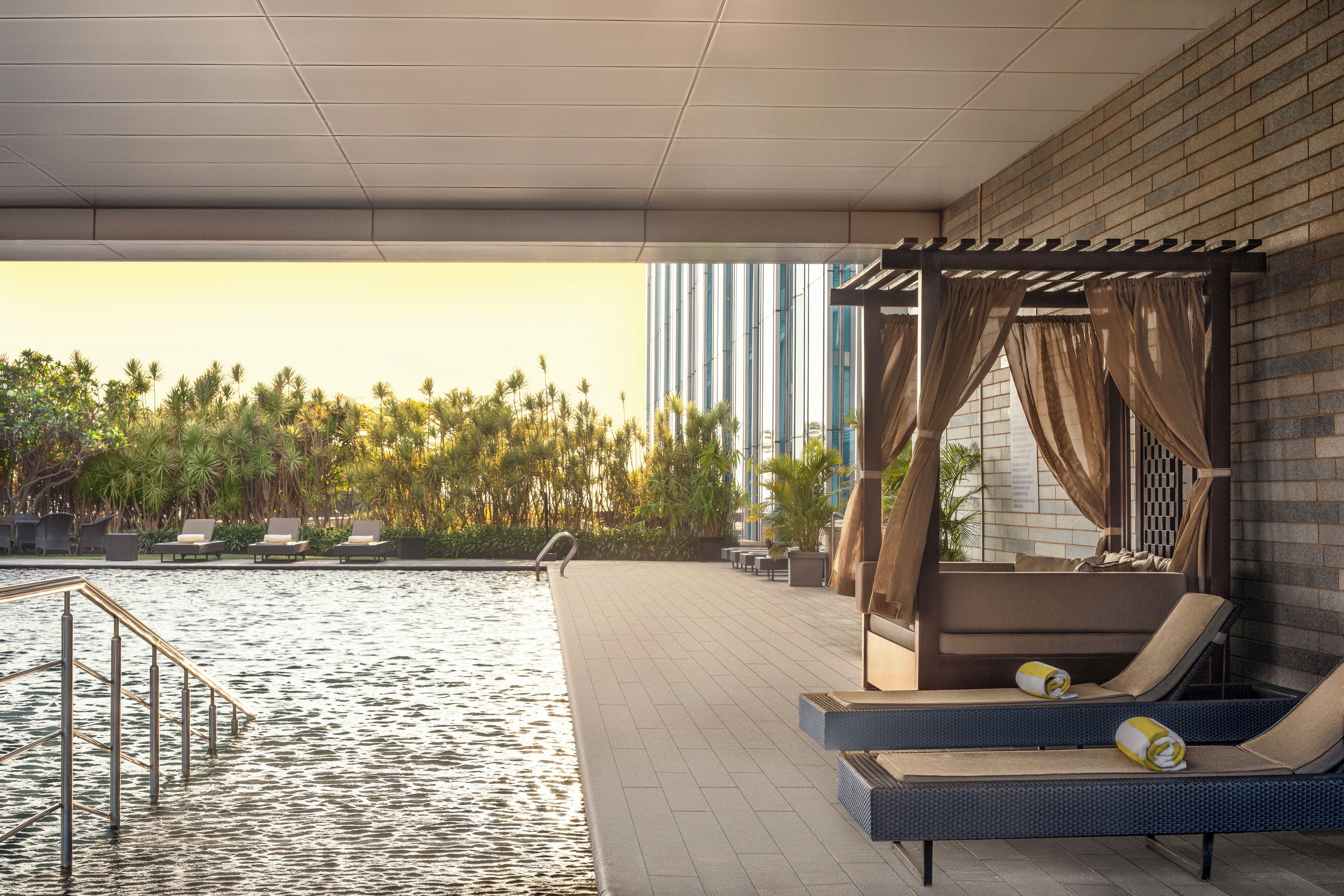 A poolside area with lounge chairs, a pergola, and a view of palm trees and buildings.