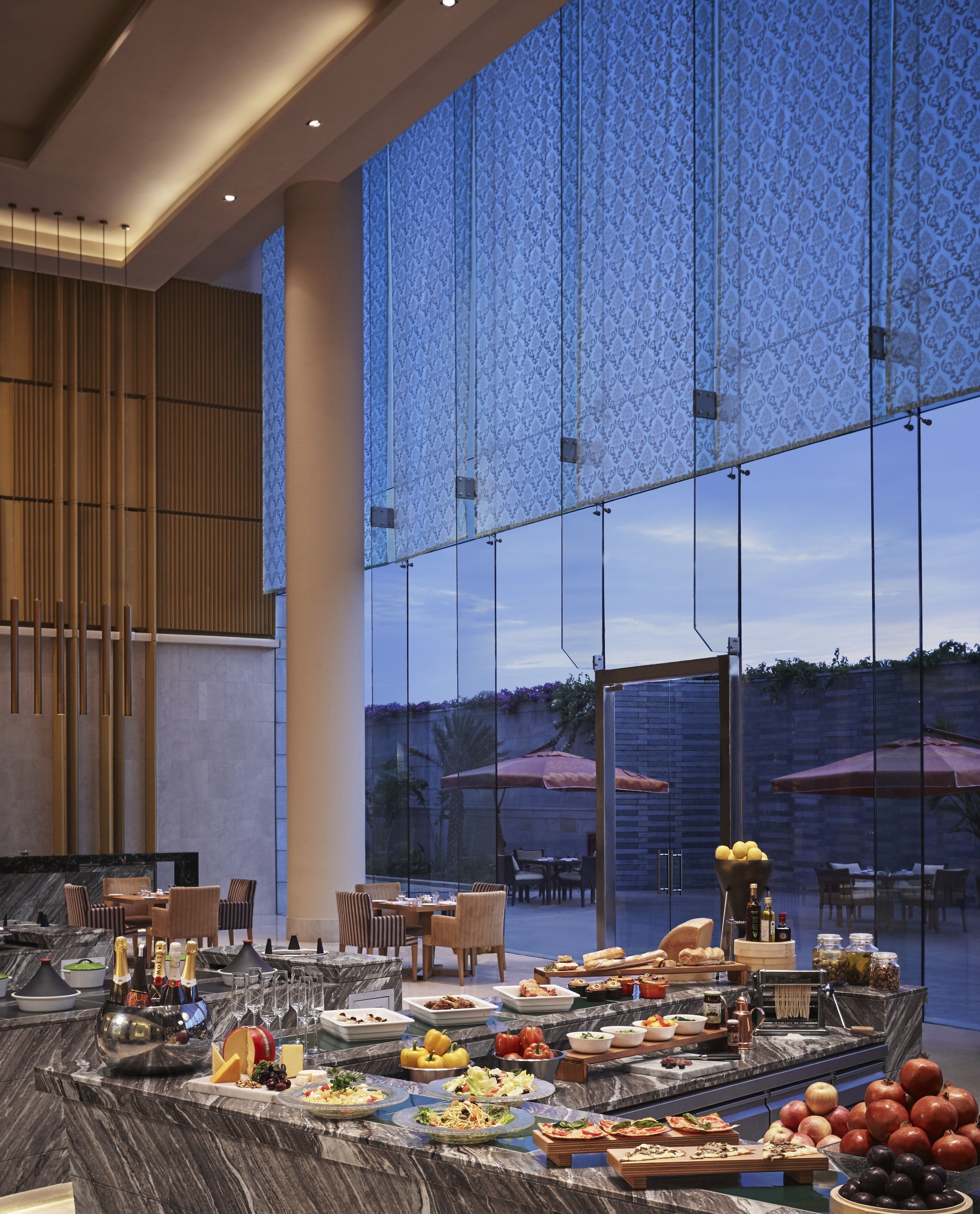 A spacious indoor dining area with a large window overlooking a garden, featuring a variety of food and drink stations.