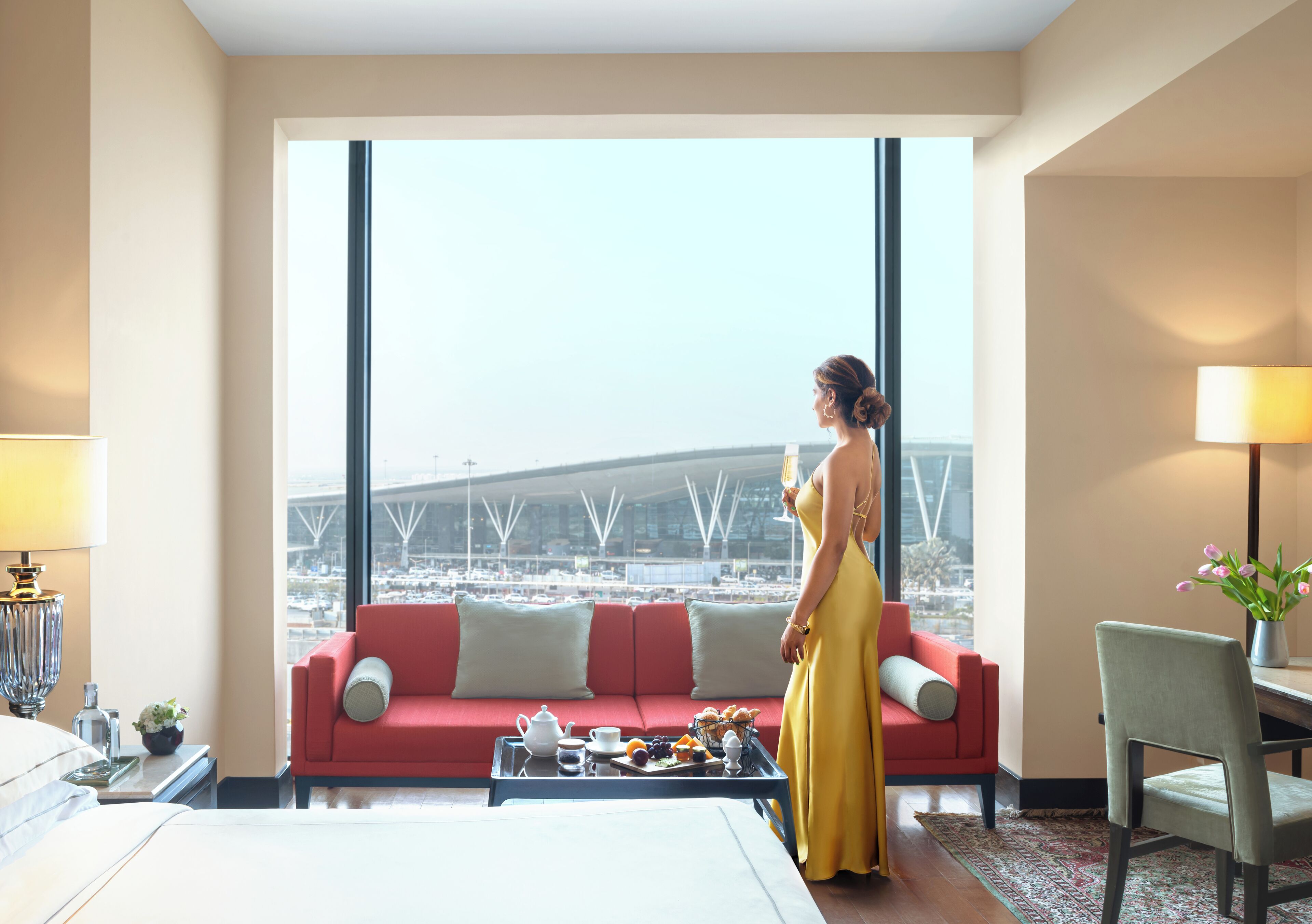 A hotel room with a large window, a red sofa, a dining table with breakfast, and a bed.