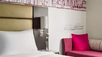 A hotel room with a bed, a nightstand, a lamp, a vase with flowers, and a couch with a vibrant pillow.